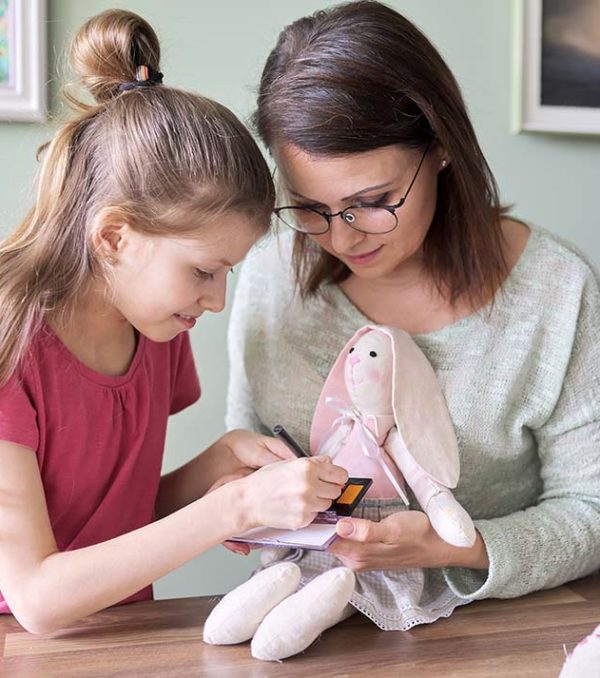 Mother and daughter child play together at home, girl and mom make make-up paints cheeks to toy handmade doll bunny sewn on sewing machine